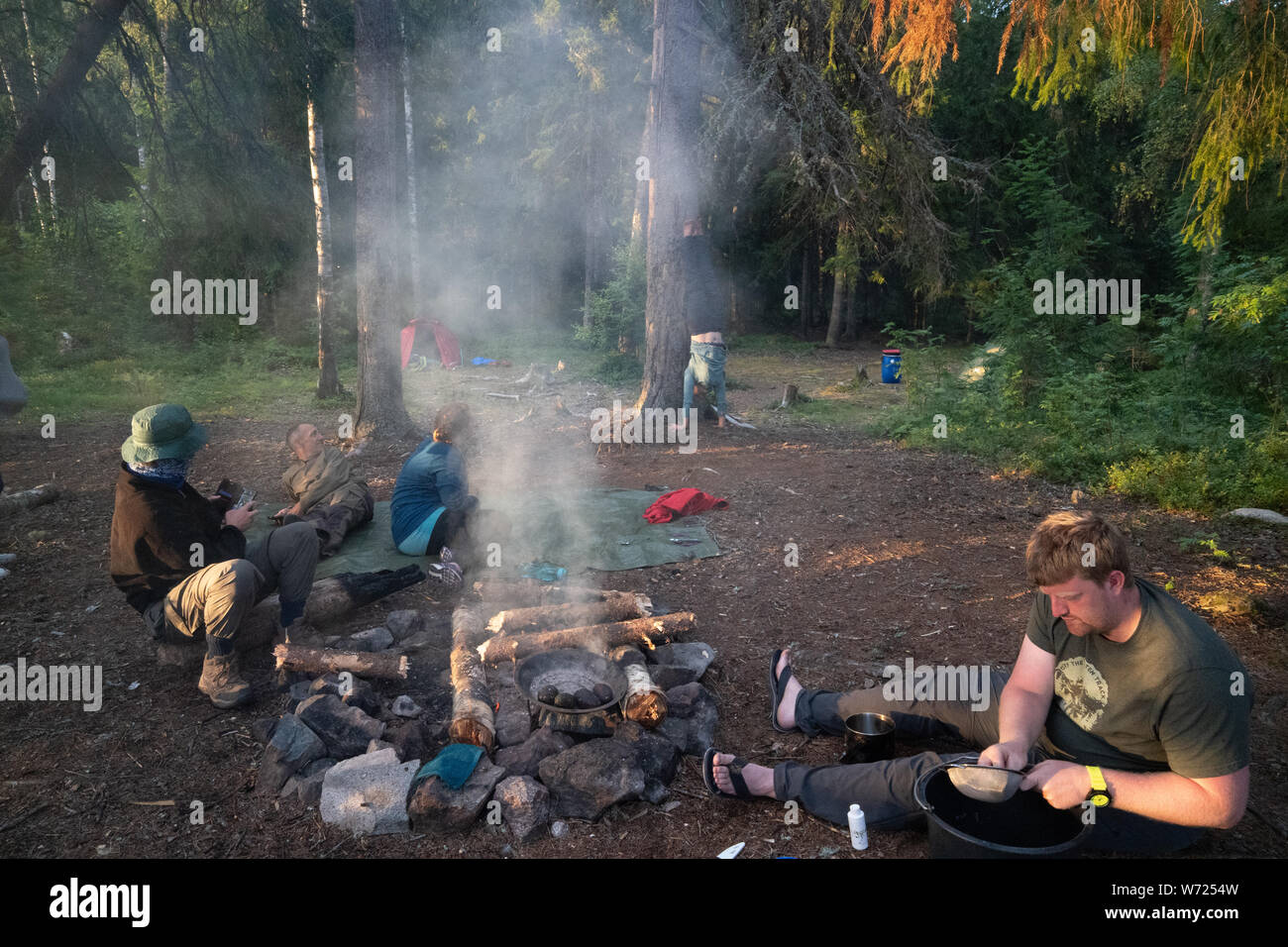 Friends wild camping in a forest sitting around a smoking camp fire Stock Photo