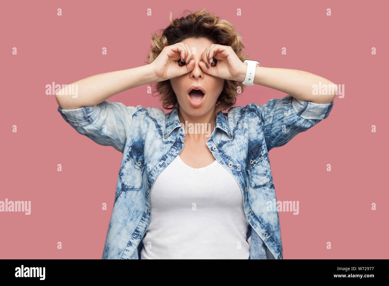 Portrait of shocked funny young woman with curly hair in blue shirt standing with binoculars gesture hands on eye and looking at camera with amazed fa Stock Photo
