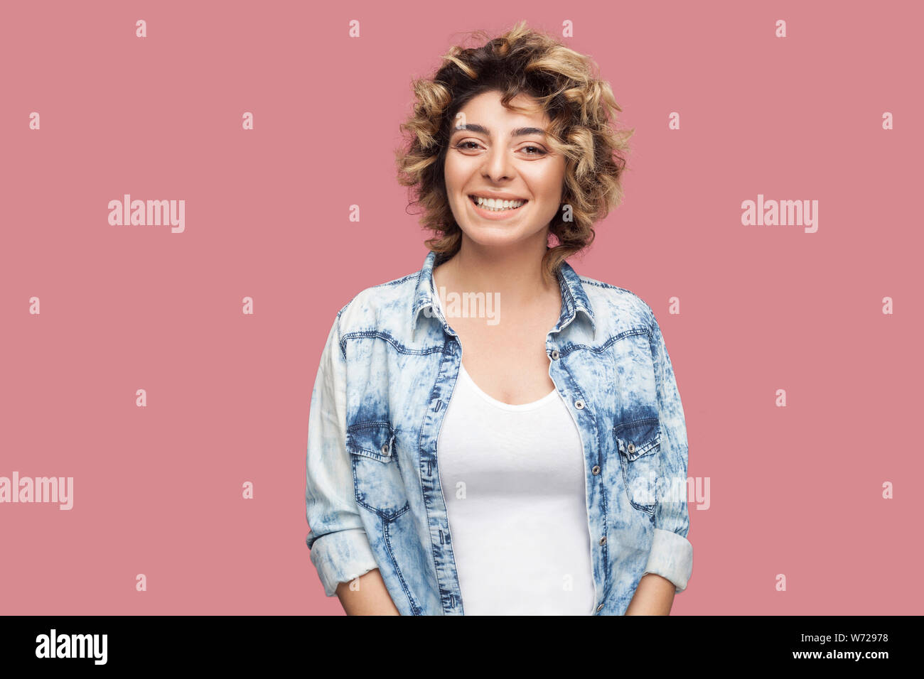 Portrait of beautiful happy young woman with makeup and curly hairstyle in casual blue shirt standing and looking at camera with toothy smile. indoor Stock Photo