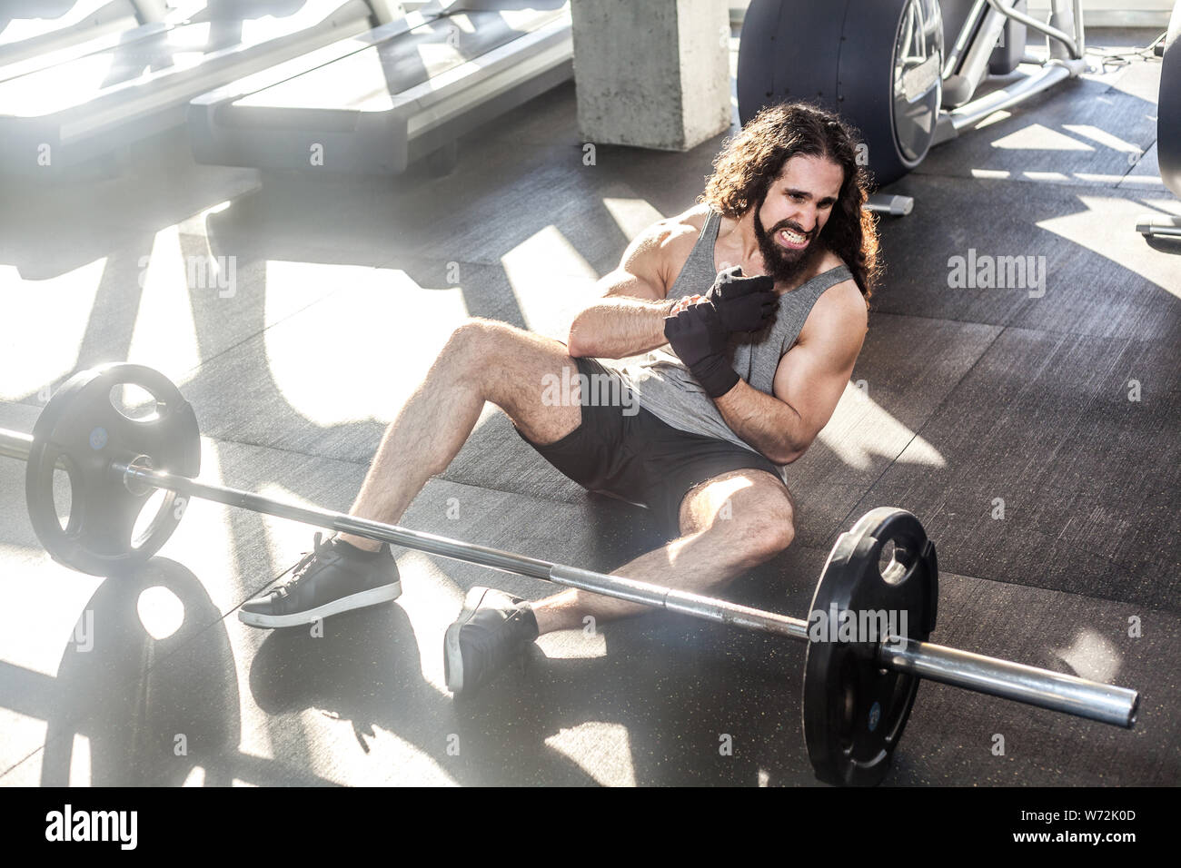 Portrait of screaming young adult man athlete with long curly hair working out in gym, sitting on floor and have strong hurt problem with wrist, spasm Stock Photo