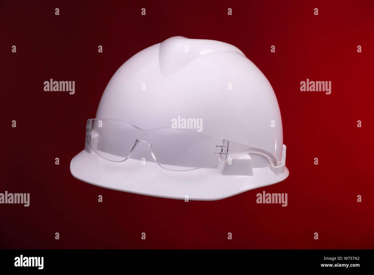 White generic protective construction helmet with a plastic transparent safety glasses on the cap against a red studio background Stock Photo