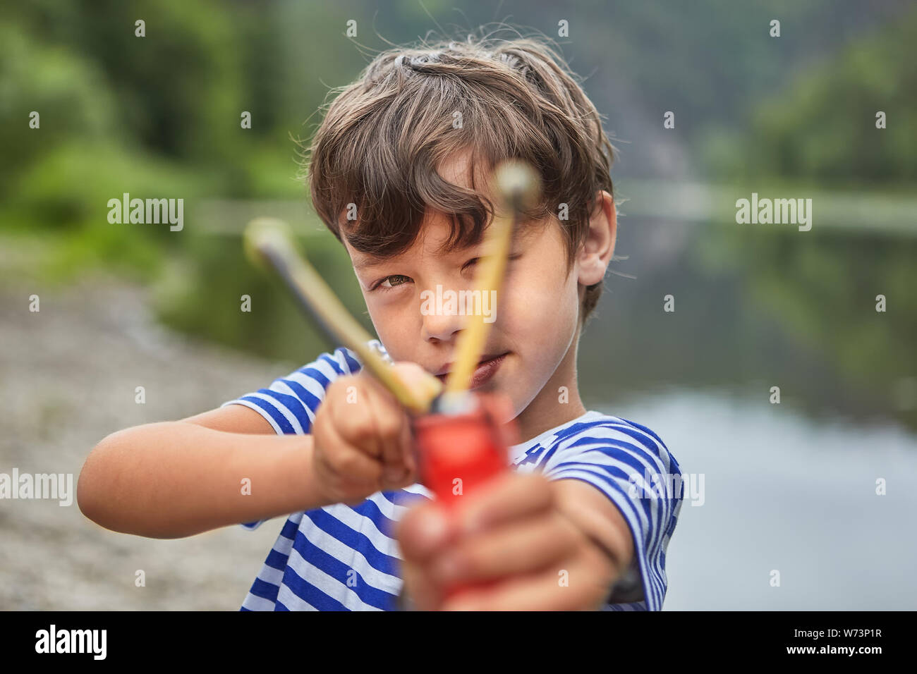 White bully boy about 8 years old is playing with slingshot in park and preparing for shot. Stock Photo