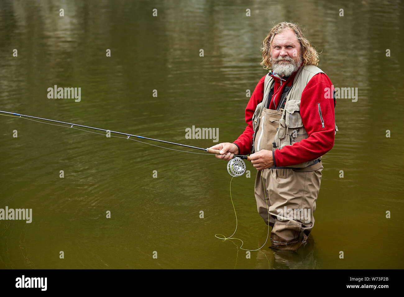 Mature bearded white man in waterproof clothing with fishing rod