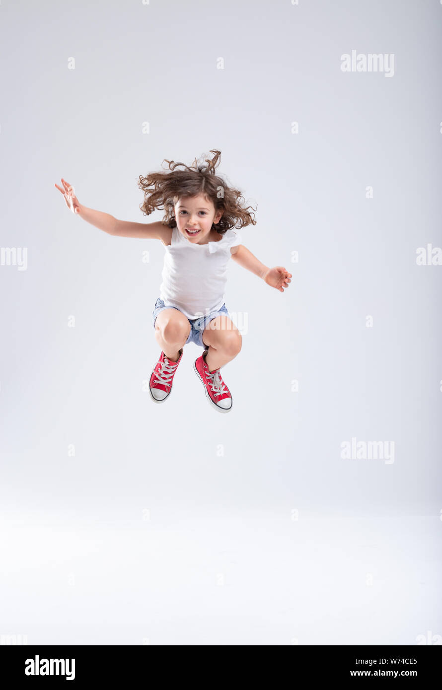 Energetic little girl jumping high into the air with bent knees and a happy smile in her red sneakers over a white background with copy space Stock Photo