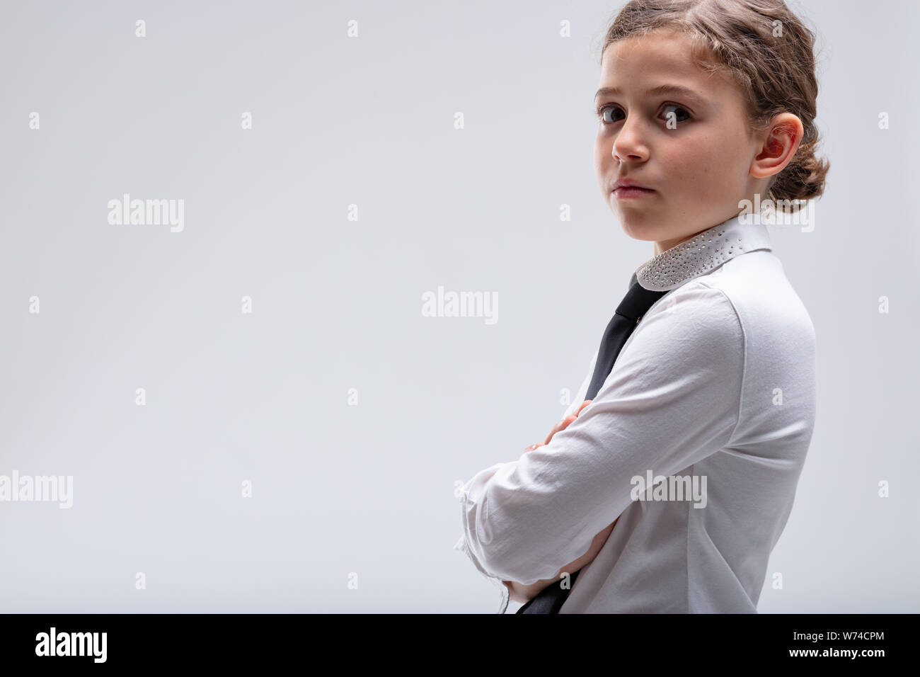 Serious young schoolgirl in uniform turning her head and looking intently at the camera with folded arms over a white background with copy space Stock Photo