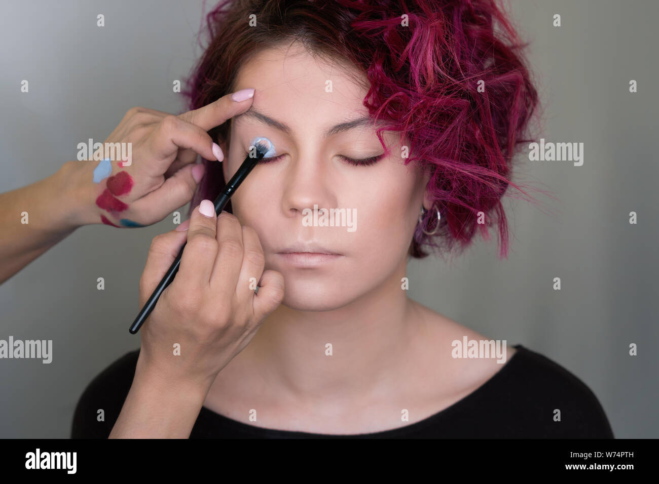 Makeup artist applies eyeshadow on the eyelid of model with special brush for makeup. Work of professional beauty artist. Bright colours. Grey backgro Stock Photo