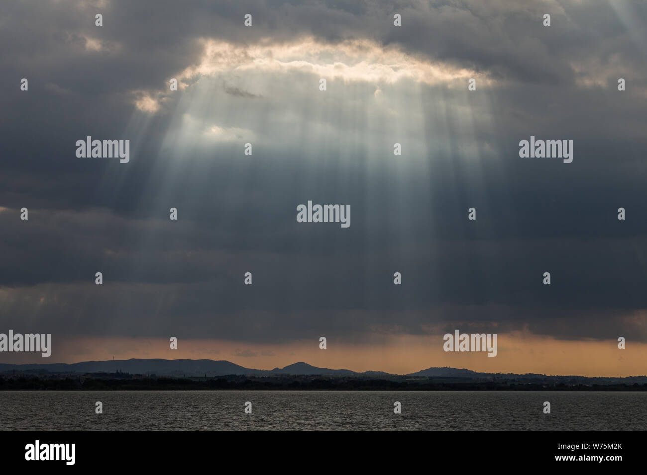 Sunrays at near sunset, with dark clouds in the background, an orange sky, and Trasimeno lake (Umbria, Italy) below Stock Photo