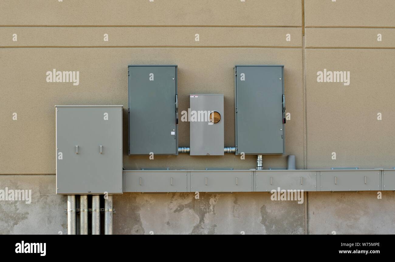Gray metal utility boxes attached to the outside wall of a building with electrical cables connecting them. Stock Photo
