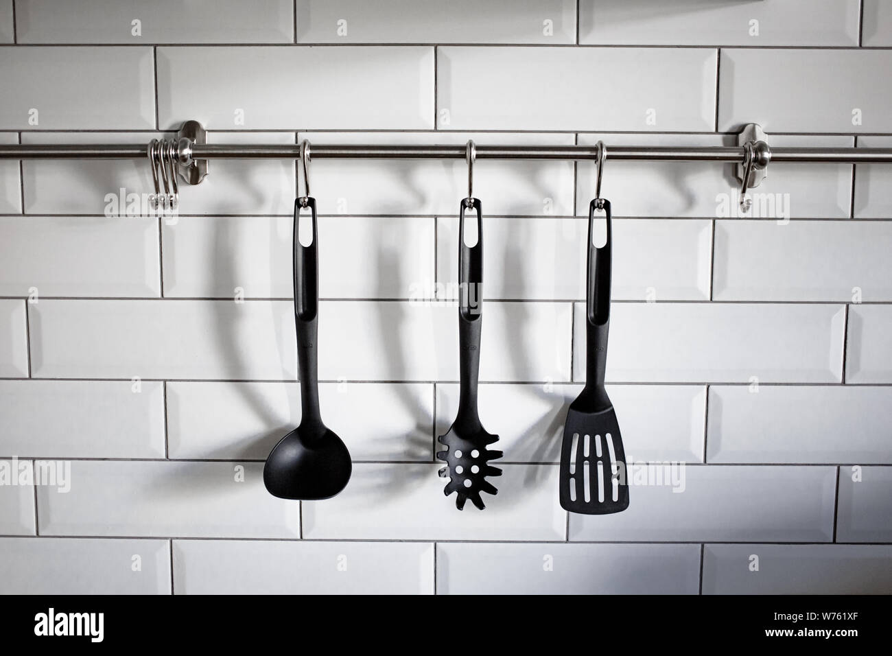 Black plastic kitchen utensil hang on white stone wall. Photo of cookware. Kitchen utensils hanging white brick background. Stock Photo