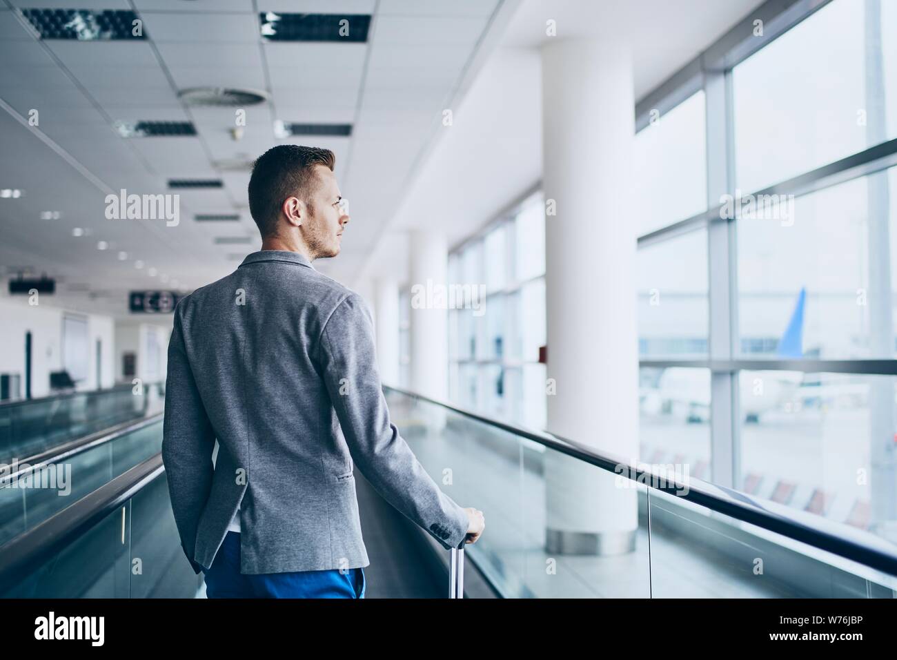 Well dressed young man travel by airplane. Businessman with luggage walking on travelator at airport. Stock Photo