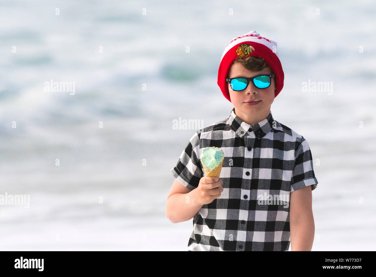 A young boy wearing reflective sunglasses, check shirt and a Chicago Blackhawks bobble hat eating an ice cream. Stock Photo