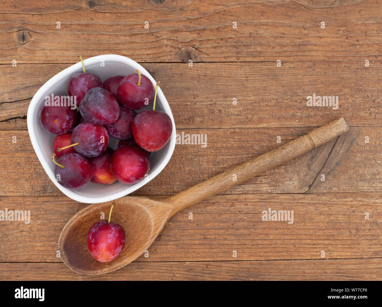 Red cherry plums in bowl, with wooden spoon, on wooden background. Stock Photo