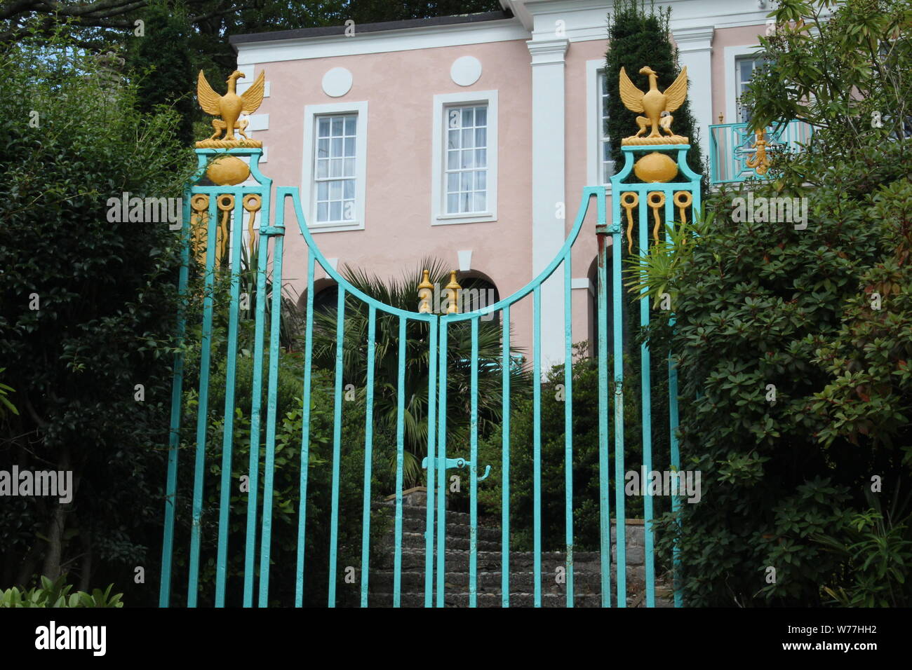 Image of a small blue gate with gold bird detailings next to some bushes, in front of a building, on a sunny day in Portmeirion Stock Photo