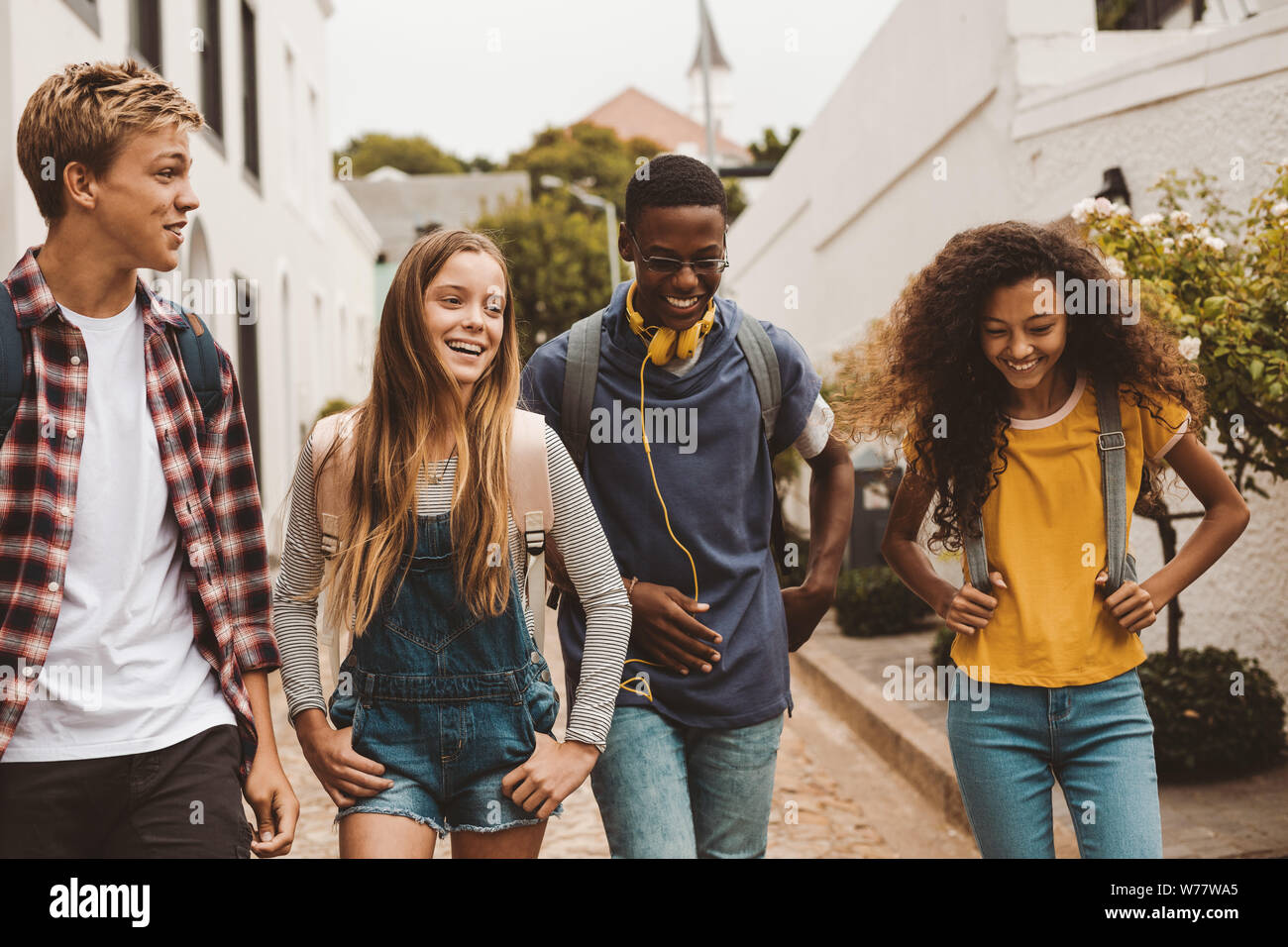Group of multiethnic college friends walking in the street. Smiling teenage friends wearing college bags walking outdoors and talking. Stock Photo