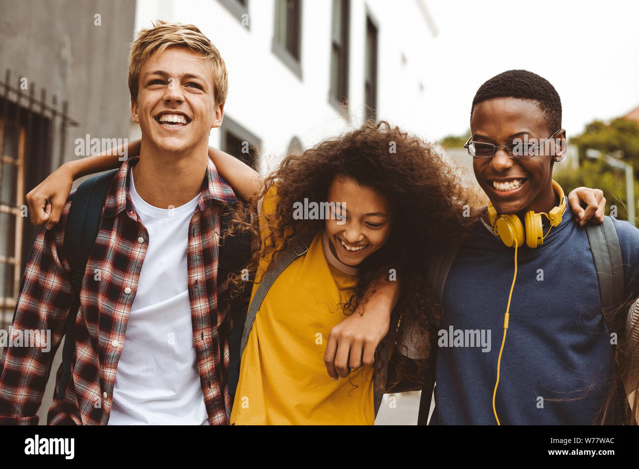 Close up of three college friends standing in the street with arms around each other. Cheerful boys and a girl wearing college bags having fun walking Stock Photo