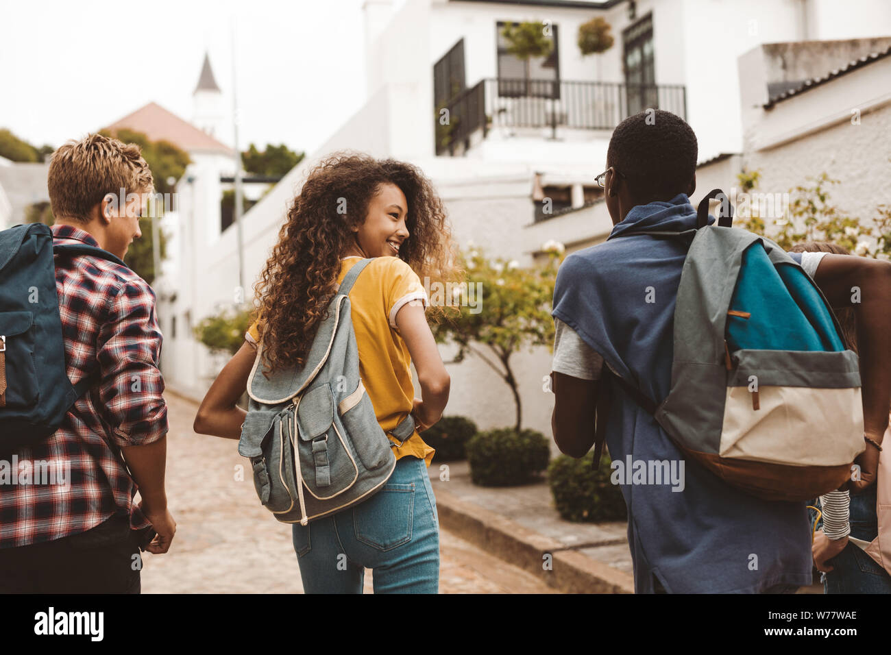 Rear view of teenage friends walking together in the street. Smiling teenage girl walking with friends wearing college bag. Stock Photo