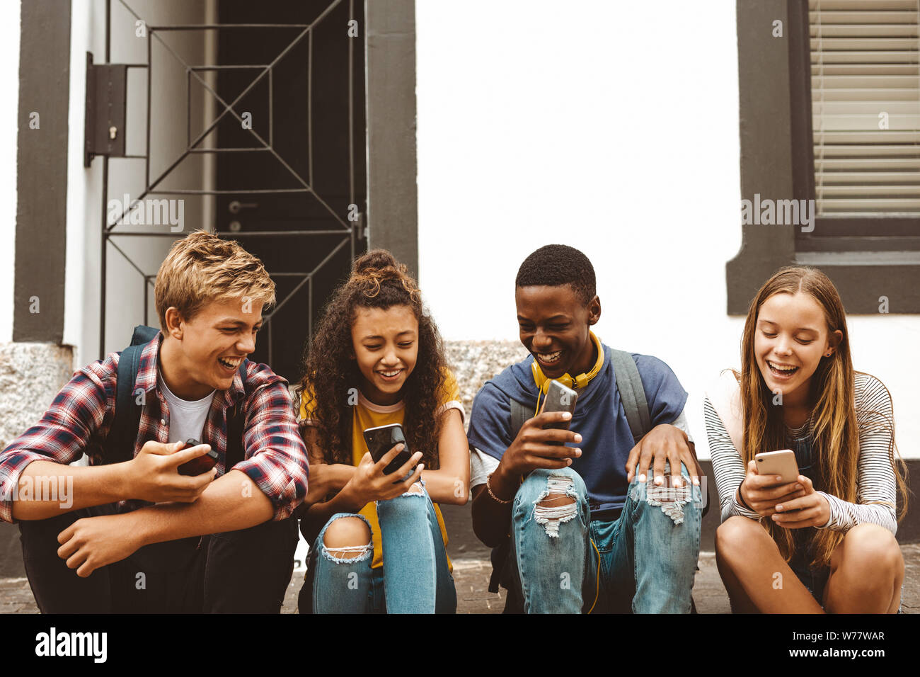 Teenage friends sitting on a pavement holding their mobile phones. Cheerful college boys and girls having fun talking sitting outdoors in a street. Stock Photo