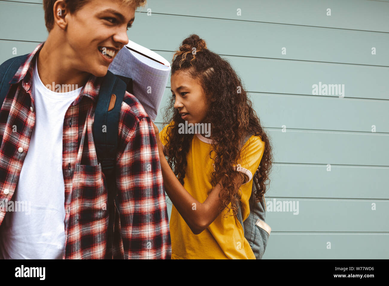 Close up of teenage friends standing outdoors wearing college bags. Girl writing in a book keeping it on the back of her friend for support. Stock Photo