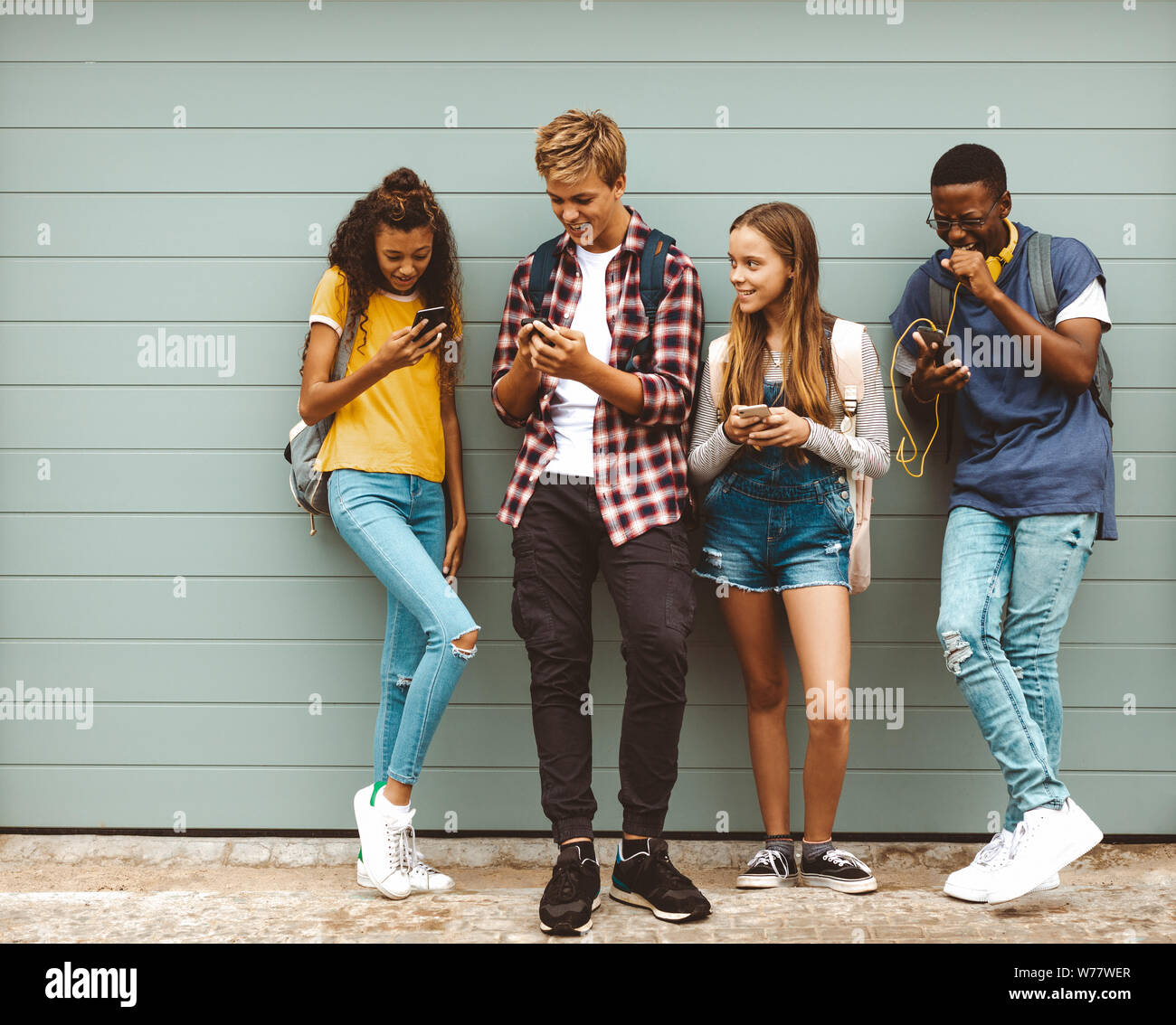 Group of multi ethnic friends standing in the street and looking at their mobile phones. Happy teenage boys and girls wearing college bags talking whi Stock Photo