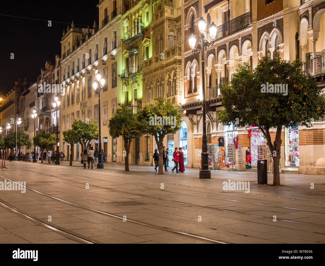 A wide street with tramtracks, shops and people waling at night in Seville old town, Spain Stock Photo