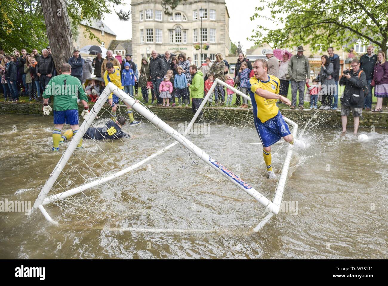 A British tradition, river football at Bourton-on-the-Water, Cotswolds August 2015. The match is held every August Bank Holiday. The game is a five-a-side football match, but in the River Windrush that runs through the town. Residents and visitors line the waters edge and bridges to get the best seats, but risk getting a soaking if the action comes too close. In 2015 British celebrithy, Griff Rhys Jones, took part during the filming for A Great British Adventure. Credit: Michael Scott/Alamy Live News Stock Photo