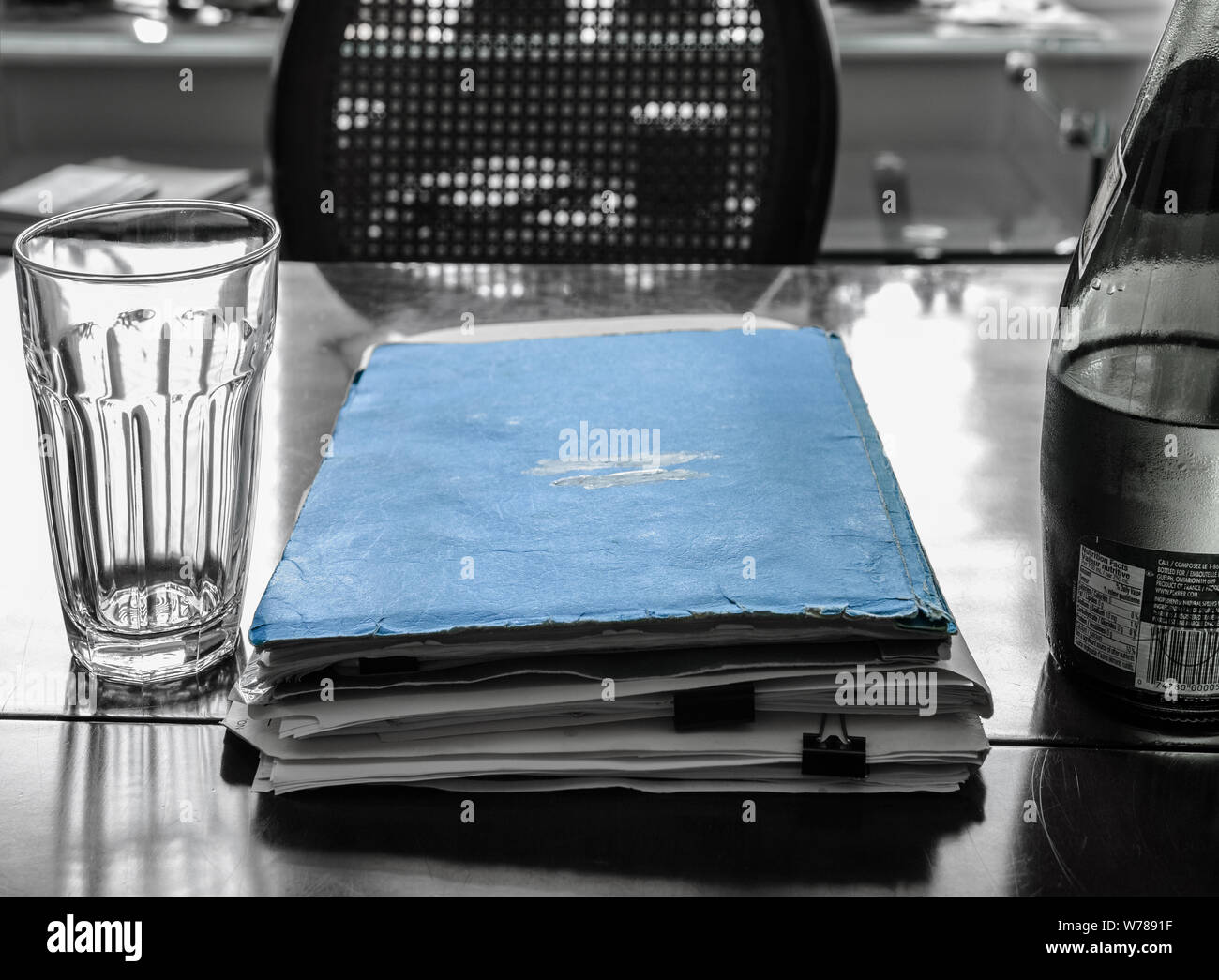 A pile of documents. The one on top has a worn out blue cover. A empty glass of water and a half full bottle of water. The chair is empty.. Stock Photo