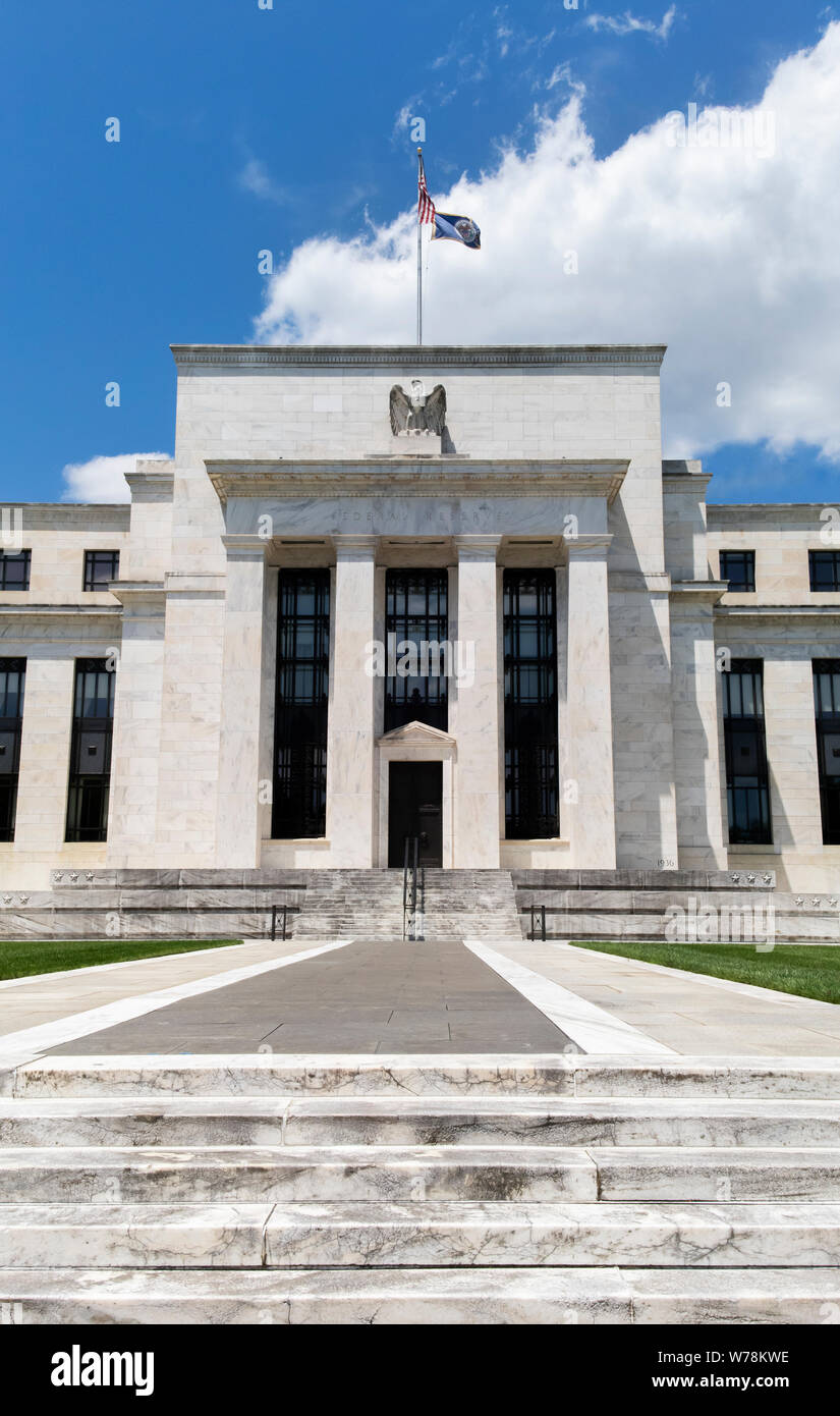Exterior view of Federal Reserve Board of Governors Building facade entrance with eagle stone statue and American flags over a blue cloudscape sky. Stock Photo