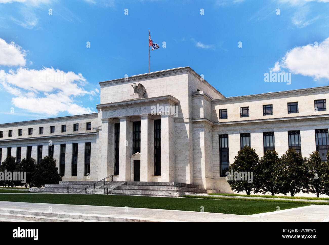 Exterior view of Federal Reserve Board of Governors Building facade entrance with eagle stone statue and American flags over a blue cloudscape sky. Stock Photo