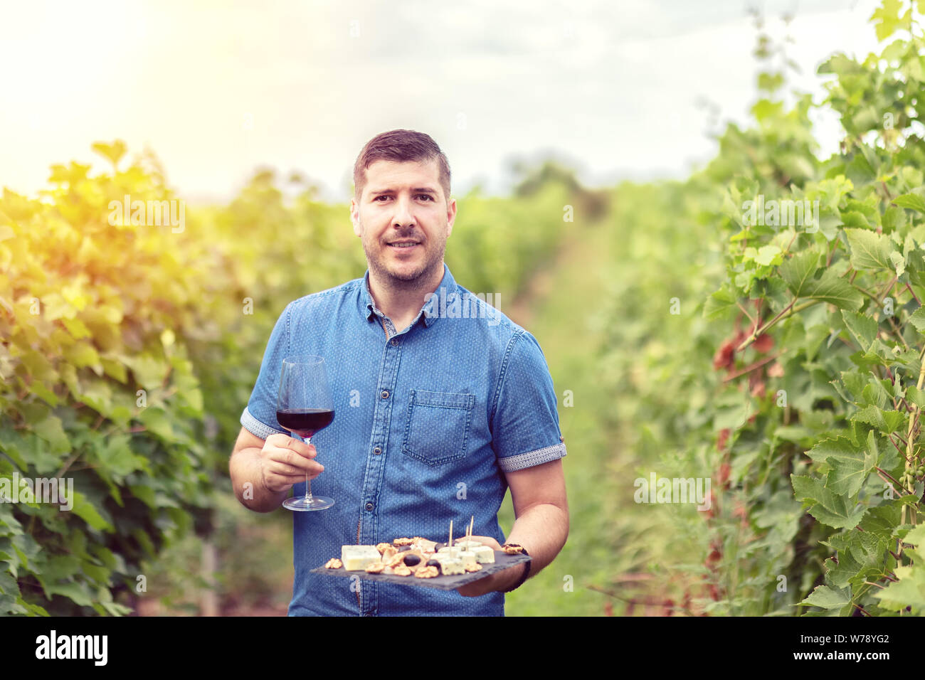 Happy successful winemaker in vineyard holding glass of red wine and board with cheese nuts and grapes – smiling man at farm house winery wine tasting Stock Photo