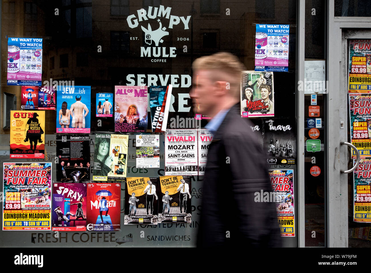 A young man walking by advertising posters for Edinburgh Fringe Festival shows on the window of an empty cafe in Edinburgh, Scotland, UK. Stock Photo