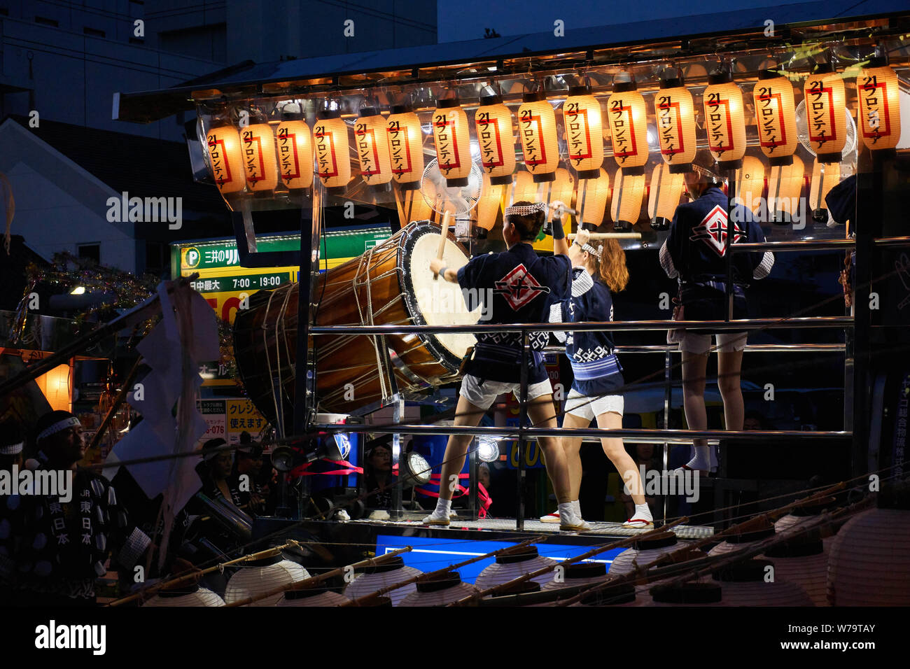 Two woman play a large taiko drum while illuminated by traditional