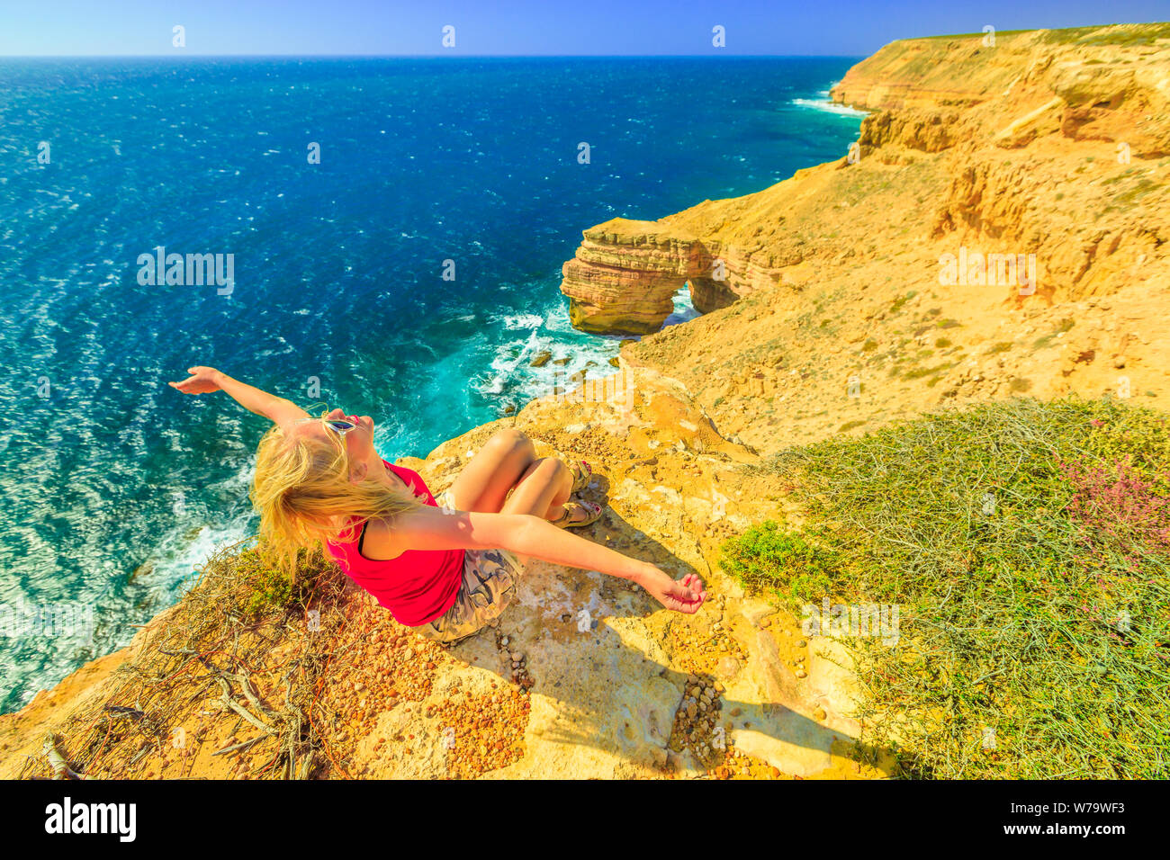 Backpacker girl with open arms at Natural Bridge in Kalbarri National Park, Western Australia. Happy woman on cliffs of Indian Ocean coastline in Stock Photo