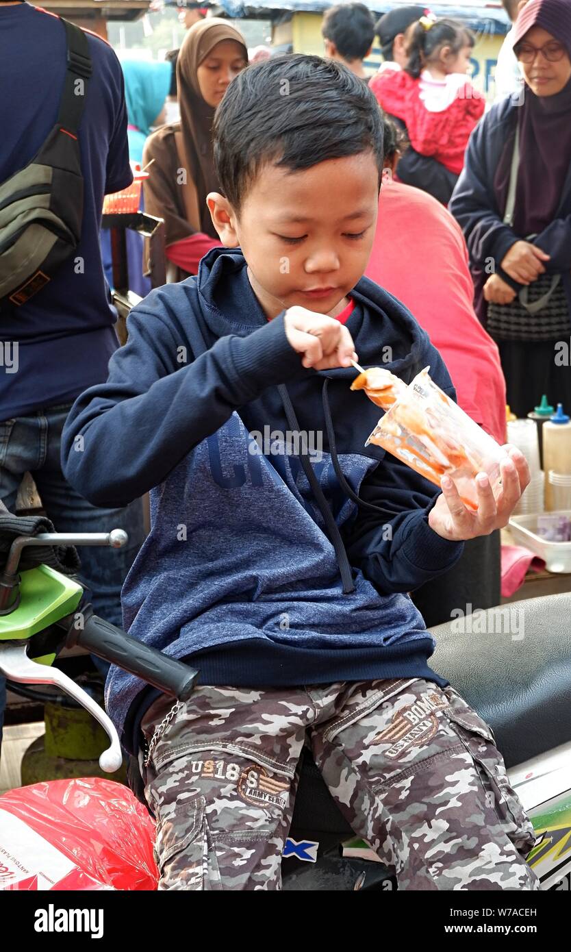 Jakarta, Indonesia - August 2019 : A boy eats a sausage with stick in front of a street food booth. Stock Photo