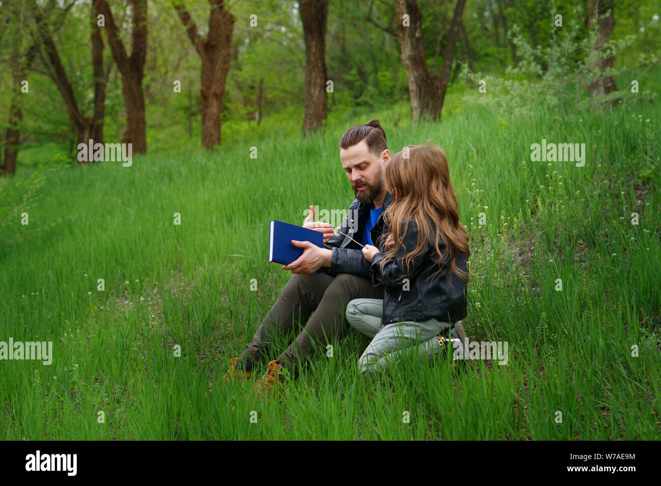 Modern stylish family walking in the park. Father and daughter are reading a book while sitting on a green lawn. Time together. Family look. Urban cas Stock Photo