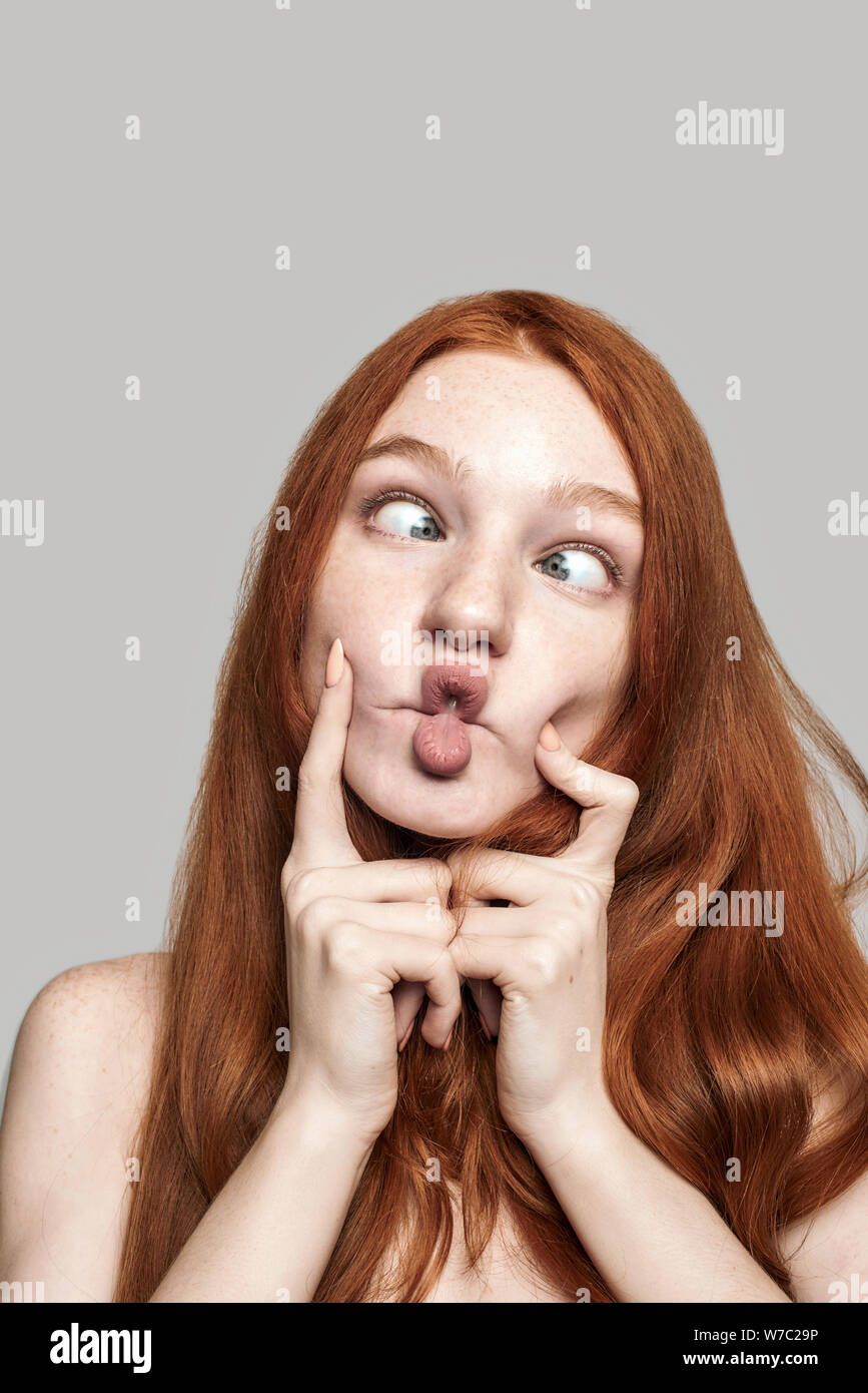 Crazy girl Close up photo of happy young redhead woman making crazy face and grimacing while standing against grey background. Human face Stock Photo