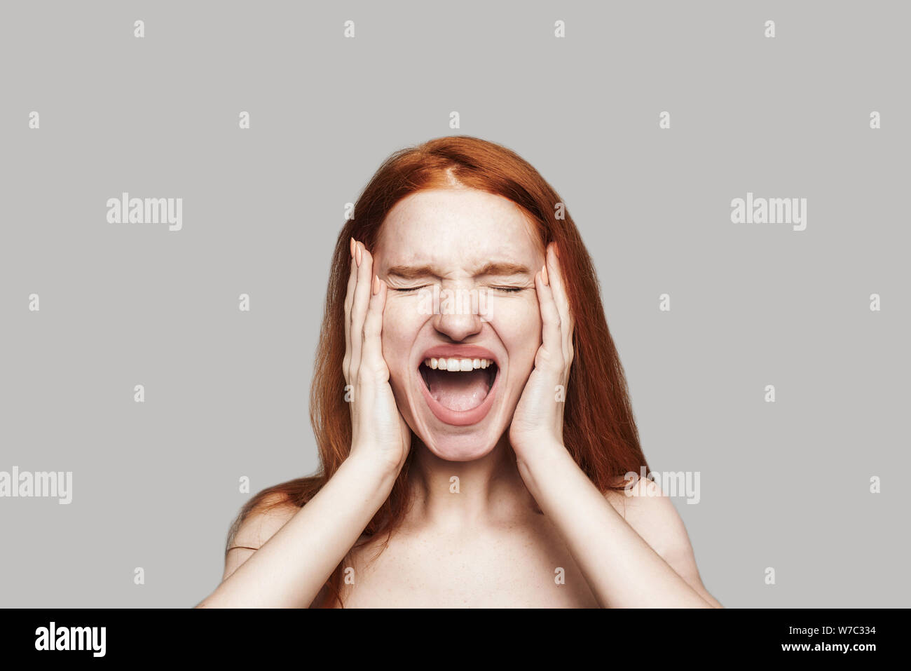 Angry stressed hysterical redhead woman with mad emotional face screaming and holding head in hands while standing against grey background. Human emotions Stock Photo
