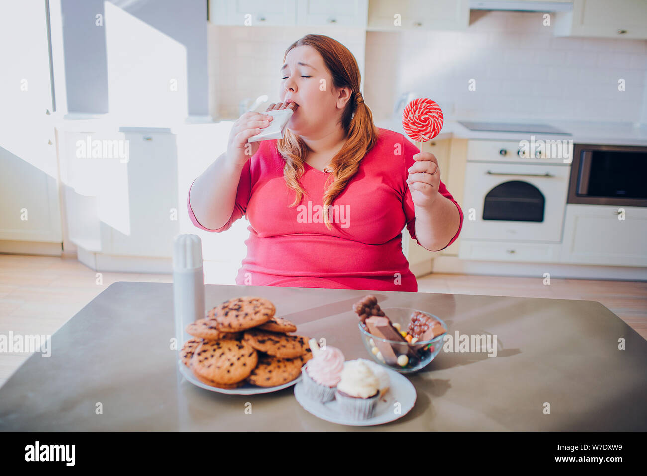 Fat young woman in kitchen sitting and eating sweet food. Biting chocolate. Cookies and cakes on table. Body positive. Daylight Stock Photo