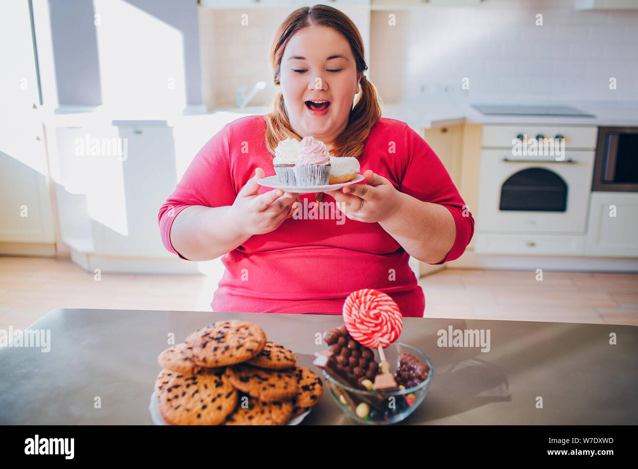 Fat young woman in kitchen sitting and eating sweet food. Happy plus size model hold cakes in hands and smile. Body positive. Daylight in kitchen Stock Photo