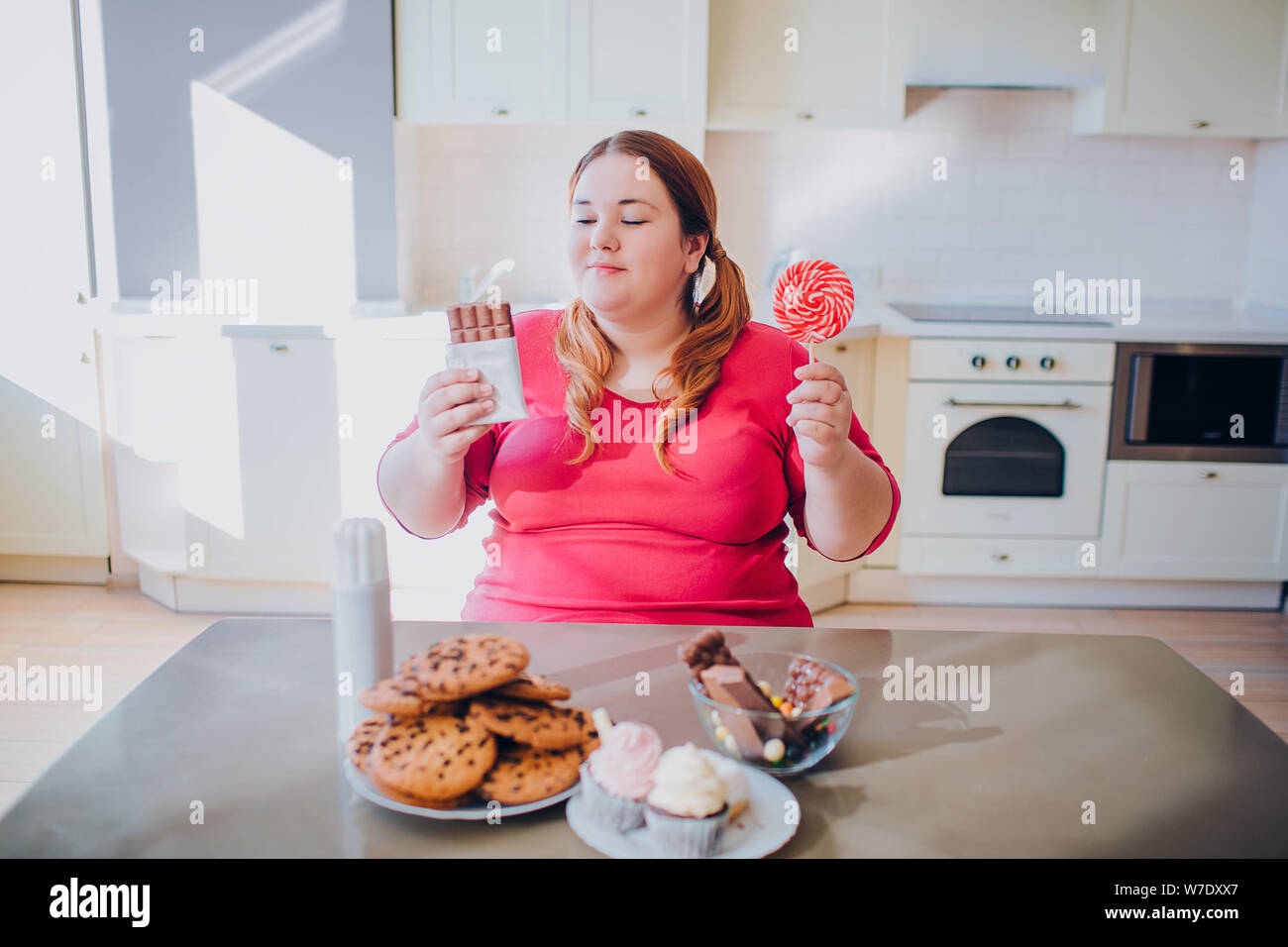 Fat young woman in kitchen sitting and eating sweet food. Cookies and cakes on table. Inside daylight. Body positive Stock Photo