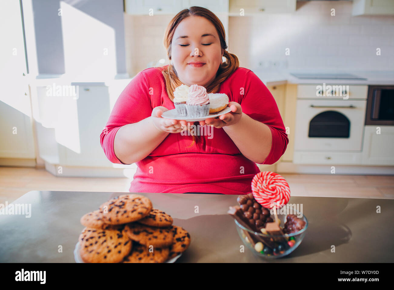 Fat young woman in kitchen sitting and eating sweet food. Plus size model hold small cakes in hands and look at them. Happy woman likes to eat. Body p Stock Photo