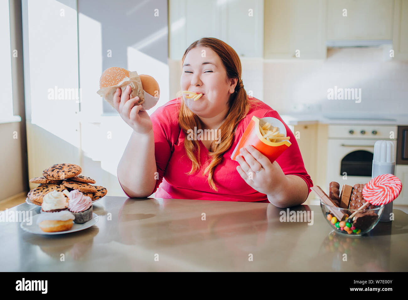 Fat young woman in kitchen sitting and eating junk food. French fries in  mouth. Looking on camera and smile. Body positive. Burgers in hands.  Unhealth Stock Photo - Alamy