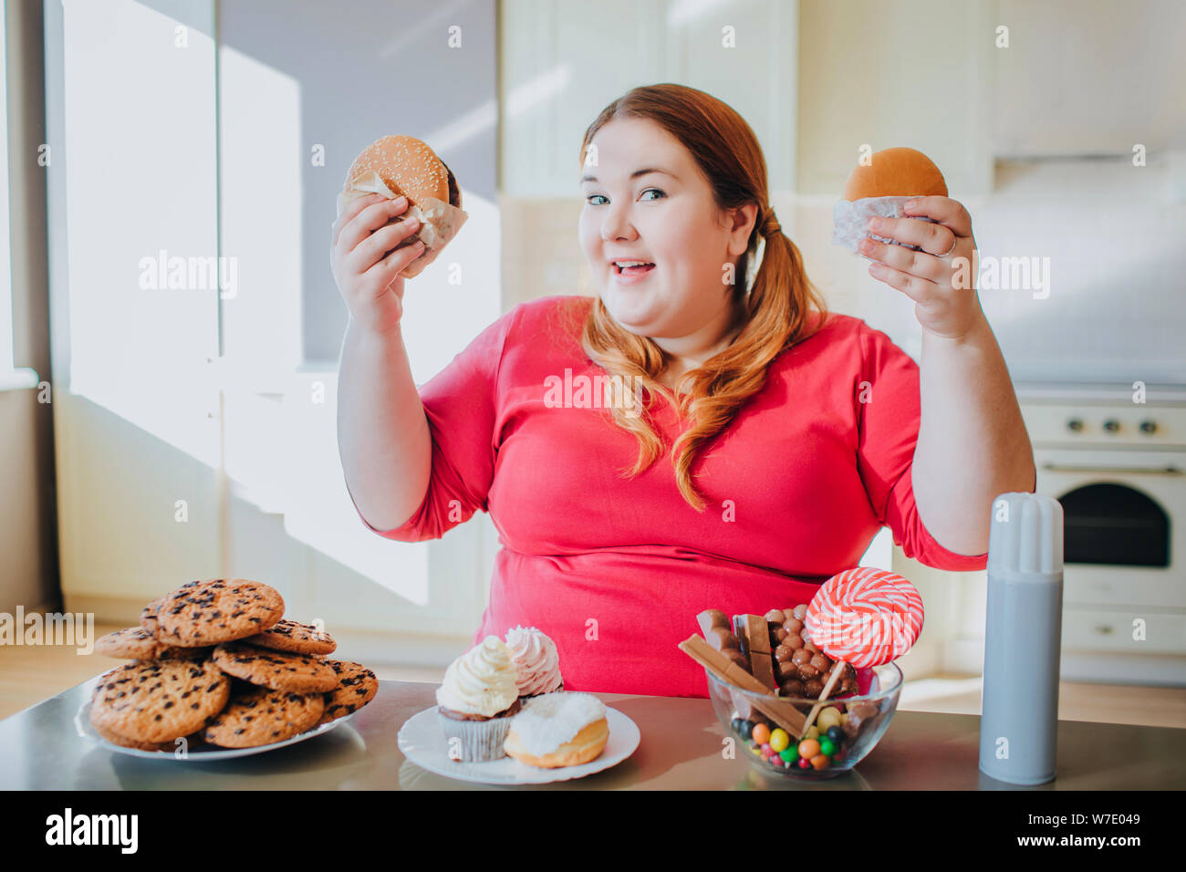 Sædvanlig prøve skulder Fat young woman in kitchen sitting and eating junk food. Holding burgers in  hands and smile to camera. Body positive. Plus size model posing. Daylight  Stock Photo - Alamy