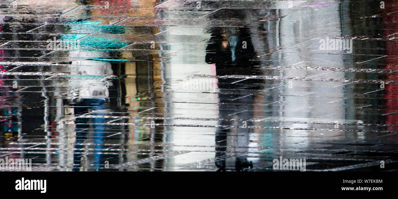 Blurry reflection shadow silhouettes of  people walking under umbrella on a rainy pedestrian city wet street, in a puddle Stock Photo