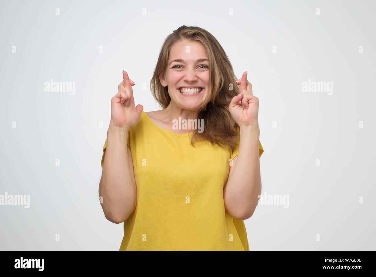 Caucasian girl in yellow shirt having excited, superstitious and naive look, keeping fingers crossed, hoping for good luck. Studio shot Stock Photo