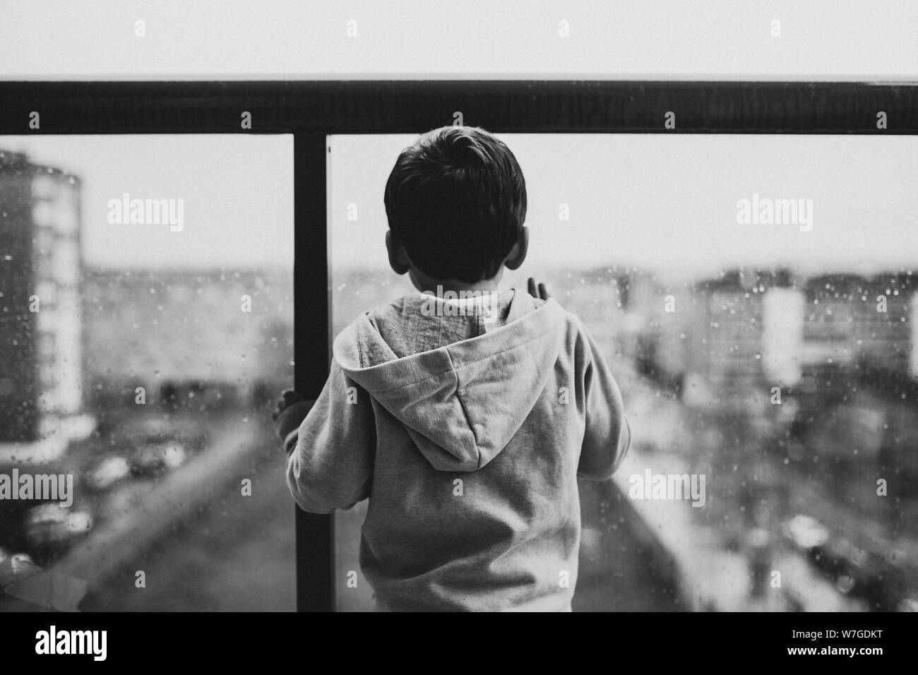 Black and white shot of a child wearing a gray hoodie standing at a balcony Stock Photo