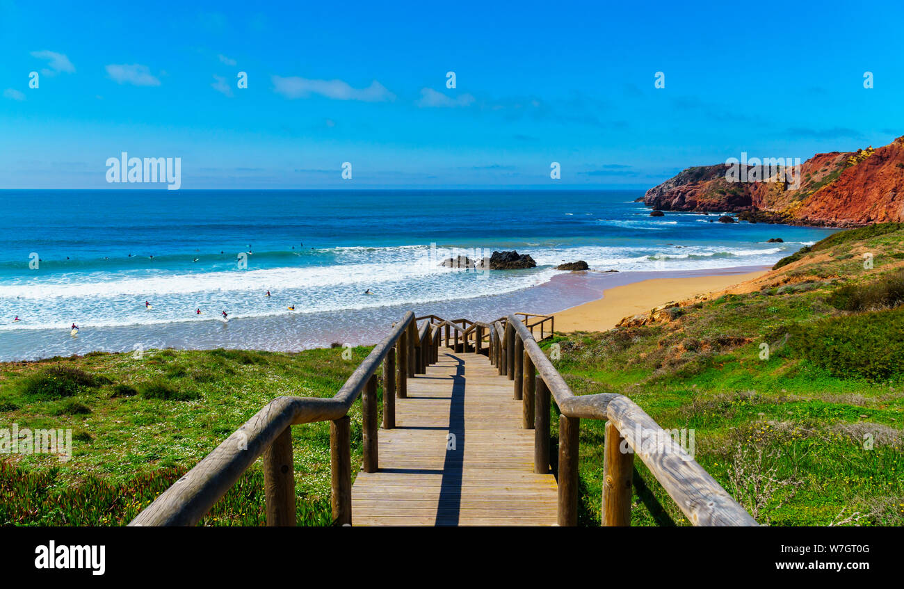 Wooden stairs with handrails leading the way through green grass down to the beach, waves, blue sky, clouds, many people on surfboards, orange cliffs Stock Photo