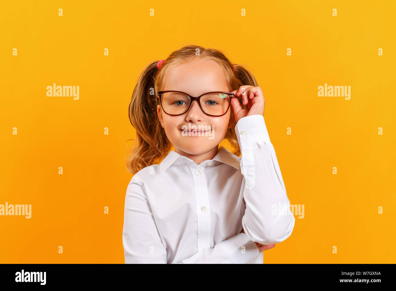 Portrait of a cute little girl on a yellow background. Child schoolgirl looks at the camera and adjusts his glasses. Education concept. Copy space. Stock Photo