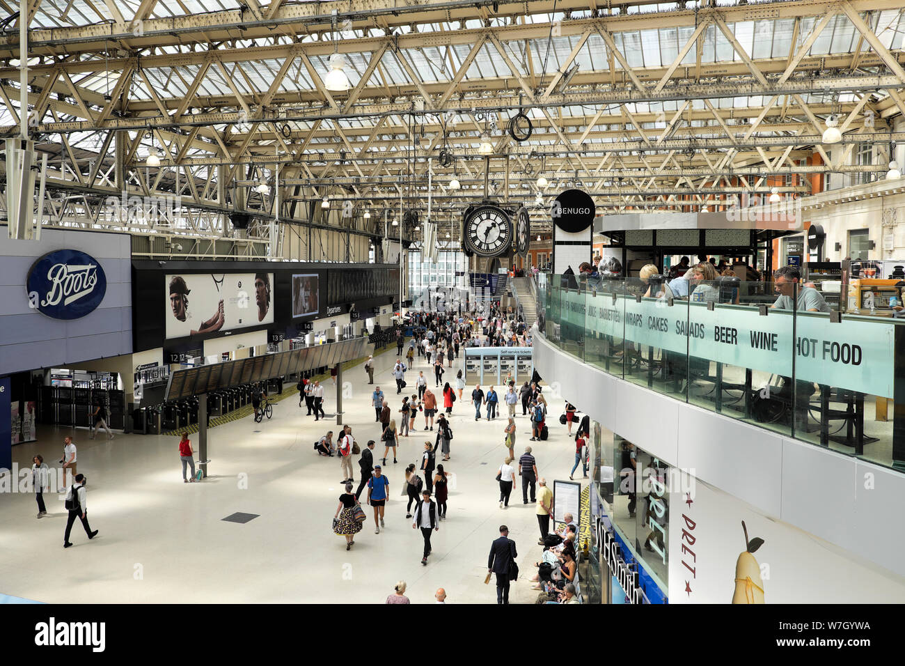 Passengers and commuters standing walking along on the concourse by Boots store under the clock