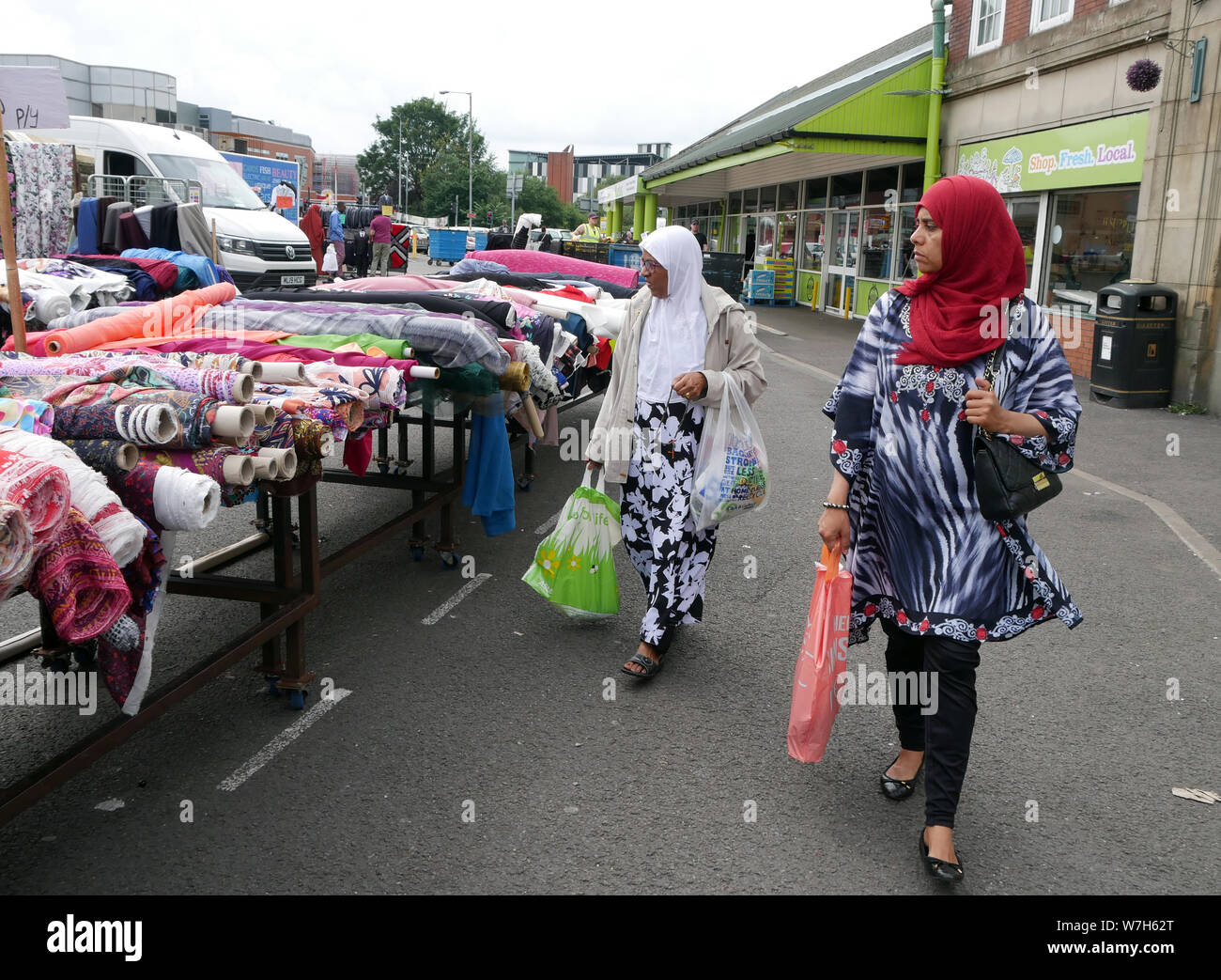 Two Asian women shoppers looking at rolls of fabric & material while shopping on Bolton outdoor market in England UK.  photo DON TONGE Stock Photo