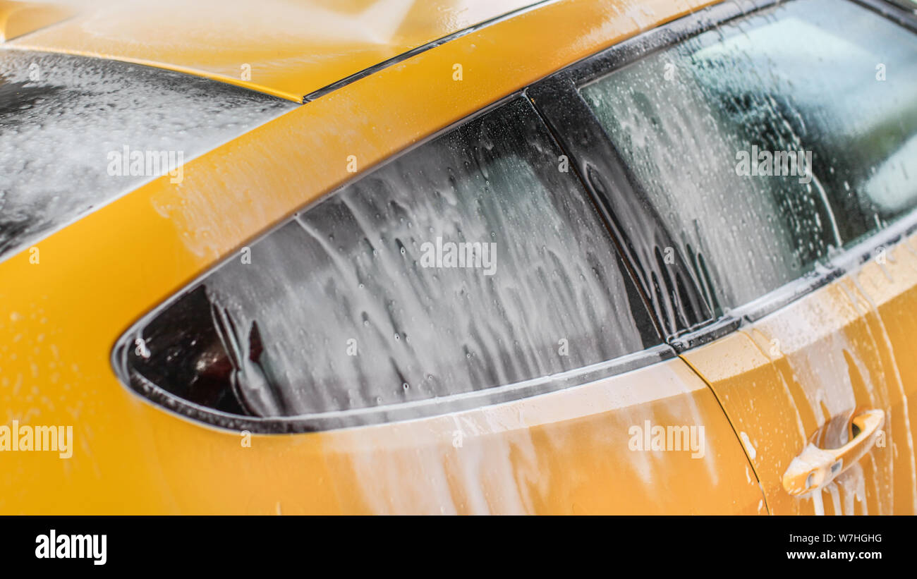 Yellow car washed in self serve carwash, shampoo spraying from hose to roof, streams of foam flowing down the window. Stock Photo